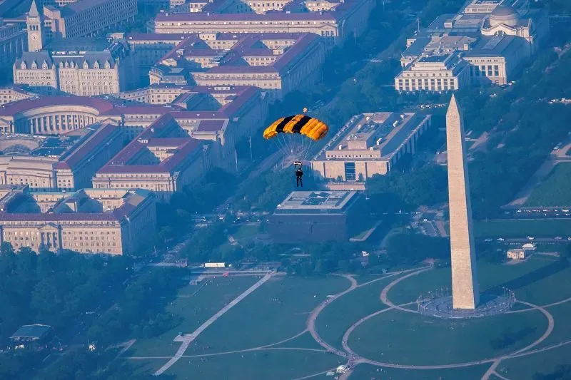مظلي ينزل بمظلة صفراء وسوداء فوق العاصمة واشنطن، مع وجود نصب واشنطن التذكاري والمباني الحكومية في الخلفية، في إطار احتفالات الجيش الأمريكي.