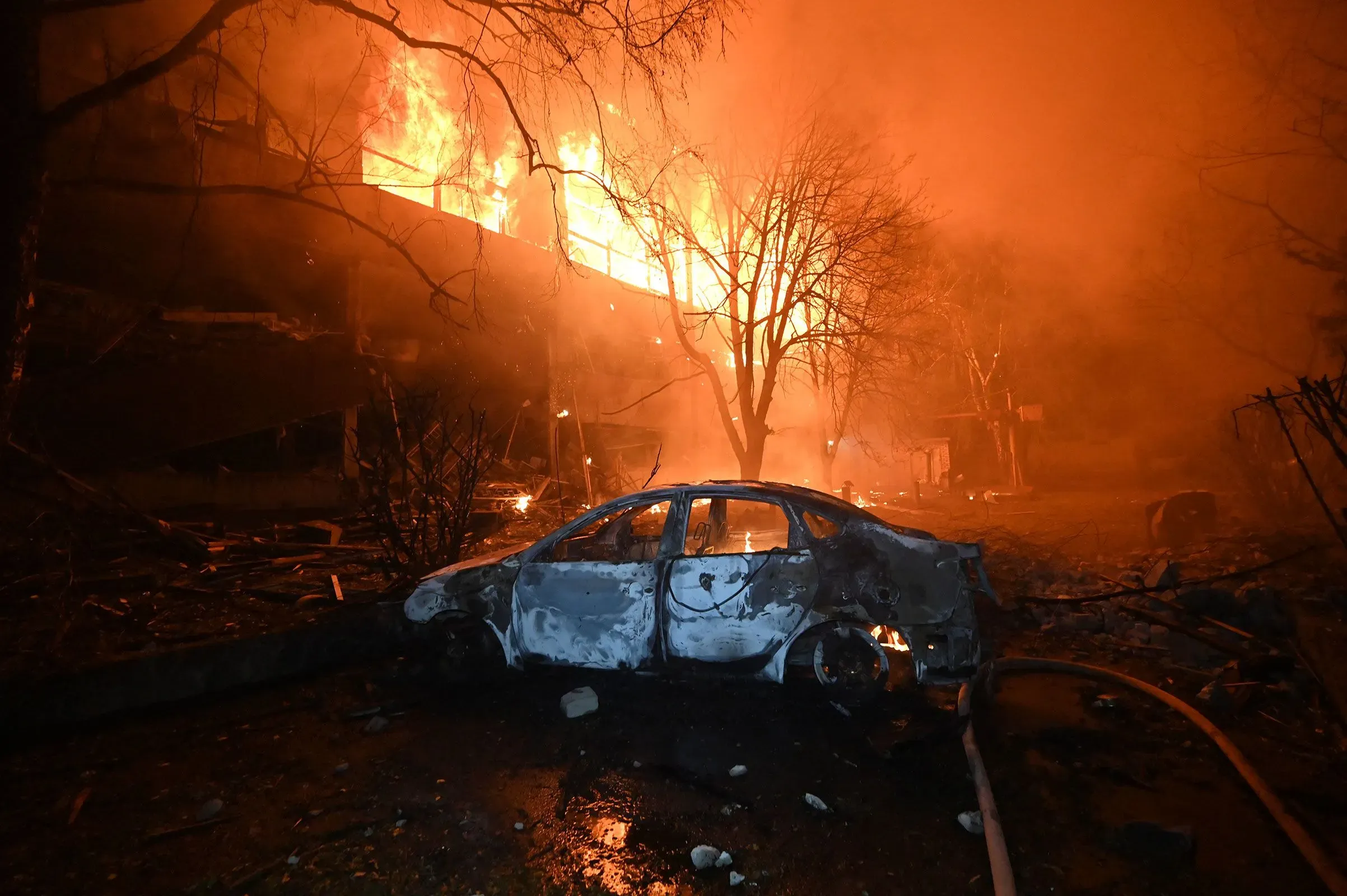 حرائق مشتعلة في مبنى خلف سيارة محترقة، تعكس آثار الحرب في أوكرانيا وتفاصيل حياة الأوكرانيين المهجرين.