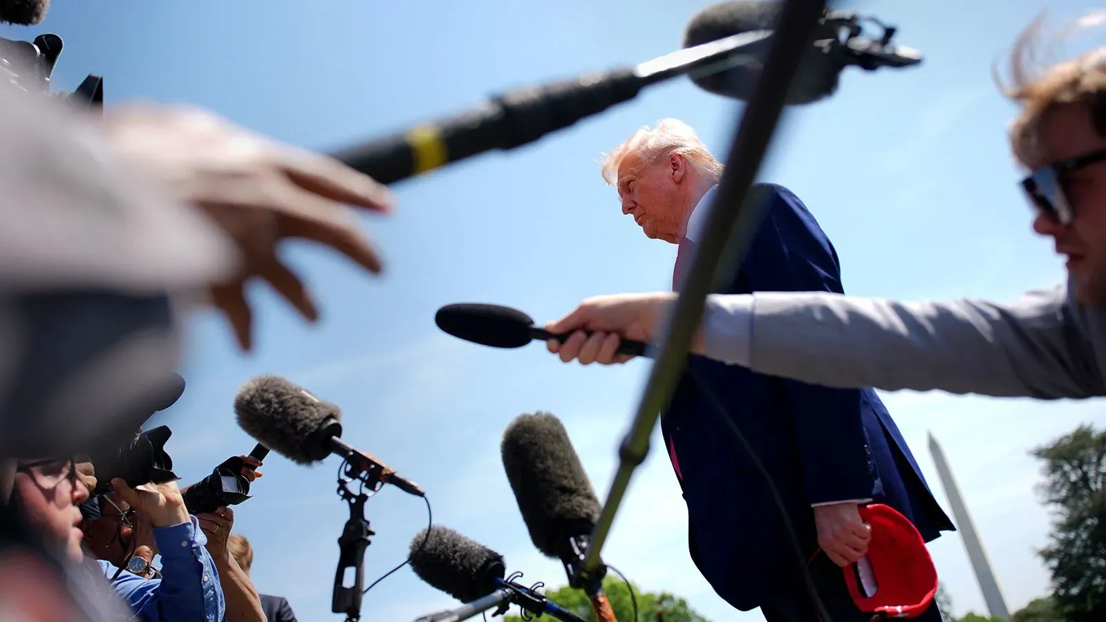 ترامب يتحدث إلى الصحفيين وسط مجموعة من الميكروفونات، مما يعكس الضغط الإعلامي الكبير الذي يواجهه خلال ولايته الثانية.