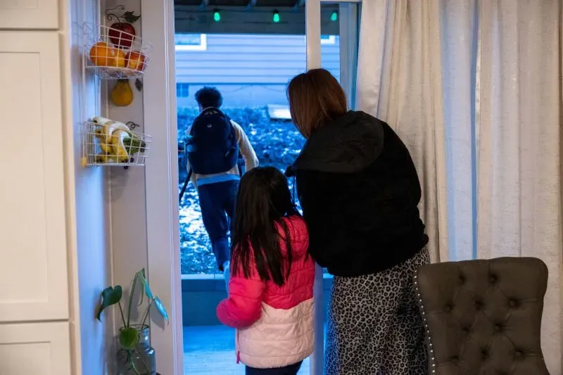 سوزانا وابنتها جيسيكا تراقبان ابنها المراهق وهو يغادر المنزل، مع لمحات من المطبخ خلفهما، مما يعكس قلق العائلة في ظل التحديات الحالية.