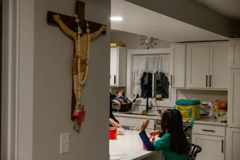 طفلة صغيرة تجلس على طاولة المطبخ، تتناول الطعام بينما يظهر صليب خشبي على الجدار، مما يعكس أجواء العائلة والتحديات التي تواجهها.