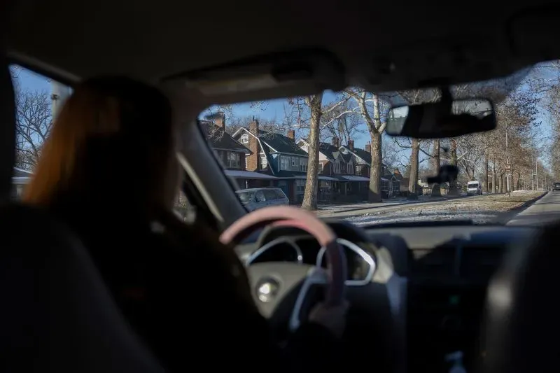 امرأة تقود السيارة، تظهر من خلف المقود، مع منازل على جانبي الطريق في يوم شتوي مشمس. تعكس الصورة شعور العائلة بالتحديات والقرارات الصعبة.