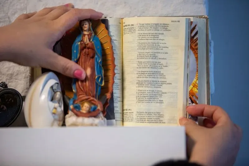 تمسك يد امرأة تمثالًا لمريم العذراء بجانب كتاب مفتوح، مما يعكس أهمية الدين والتراث في حياة عائلتها.