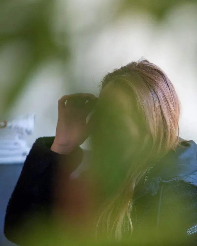 امرأة ذات شعر بني طويل، تضع يدها على وجهها، تظهر من خلف أوراق نباتية، تعكس شعور القلق والخوف من التغيرات السياسية.