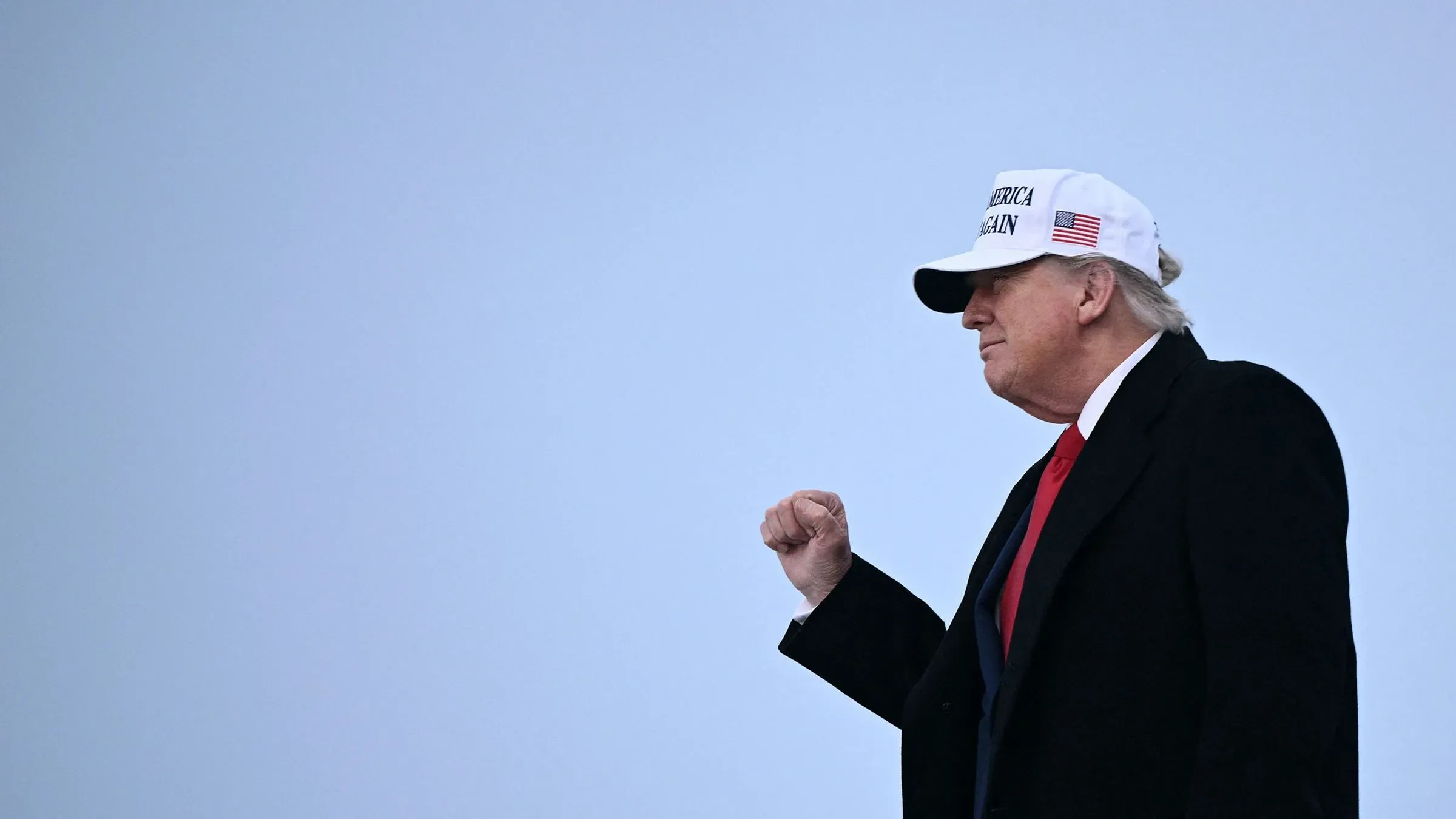 ترامب يسير مرتديًا قبعة تحمل شعار "اجعل أمريكا عظيمة مرة أخرى"، مع تعبير جاد، في خلفية رمادية.