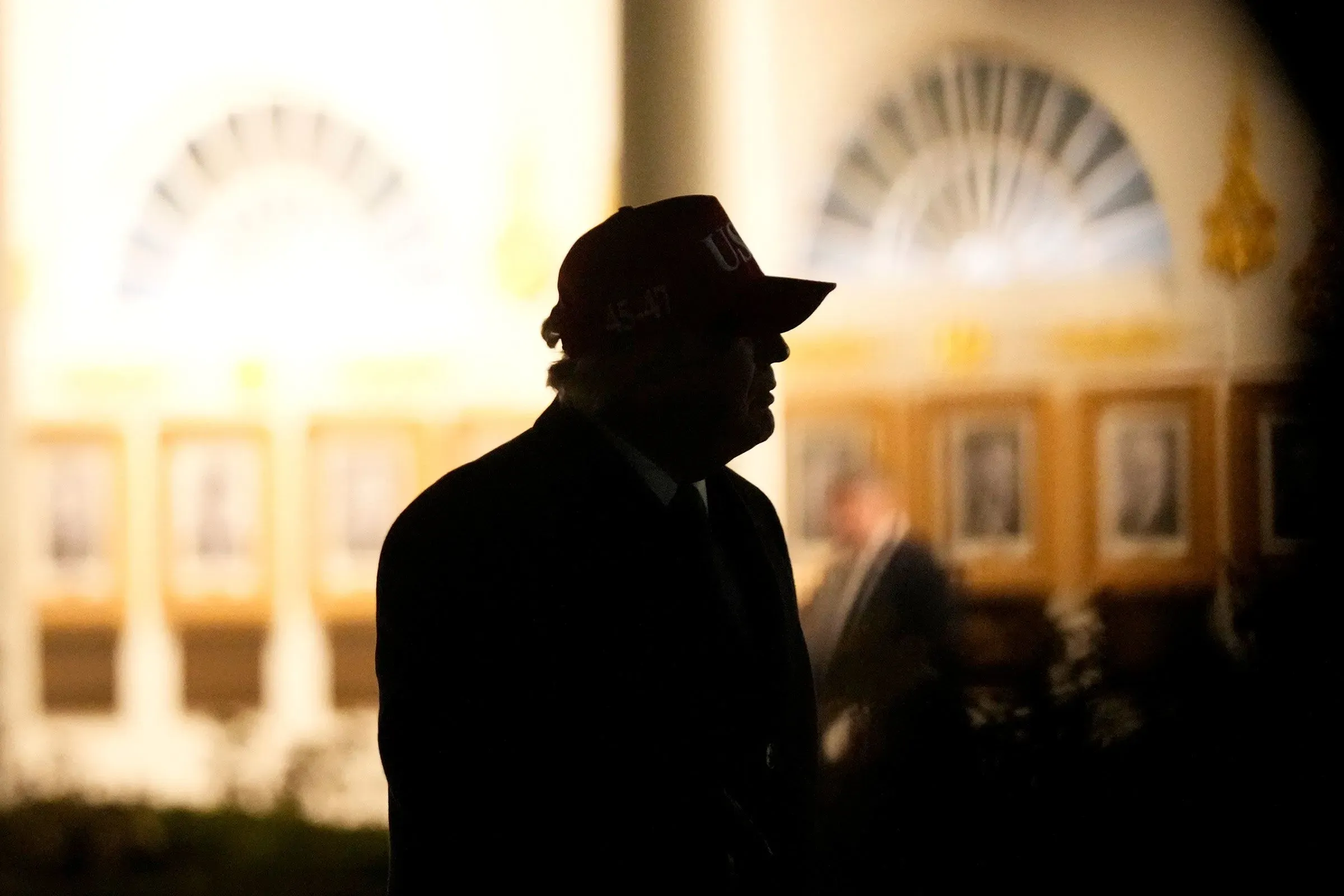 صورة لترامب في الظل، يرتدي قبعة حمراء، مع خلفية مضاءة تظهر تفاصيل معمارية، تعكس سياق السياسة الخارجية الأمريكية.