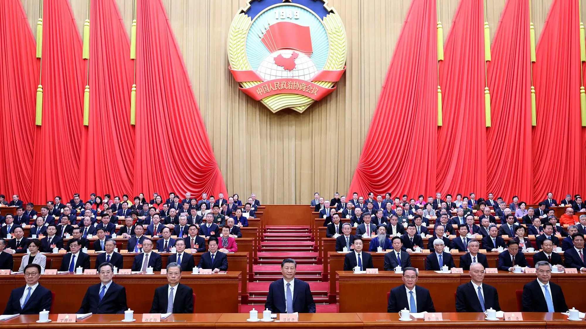 اجتماع سياسي كبير في الصين مع شي جين بينغ في المقدمة، حيث يجلس المسؤولون في قاعة مزينة بالأعلام الحمراء، مع التركيز على تعزيز الابتكار والنمو الاقتصادي.