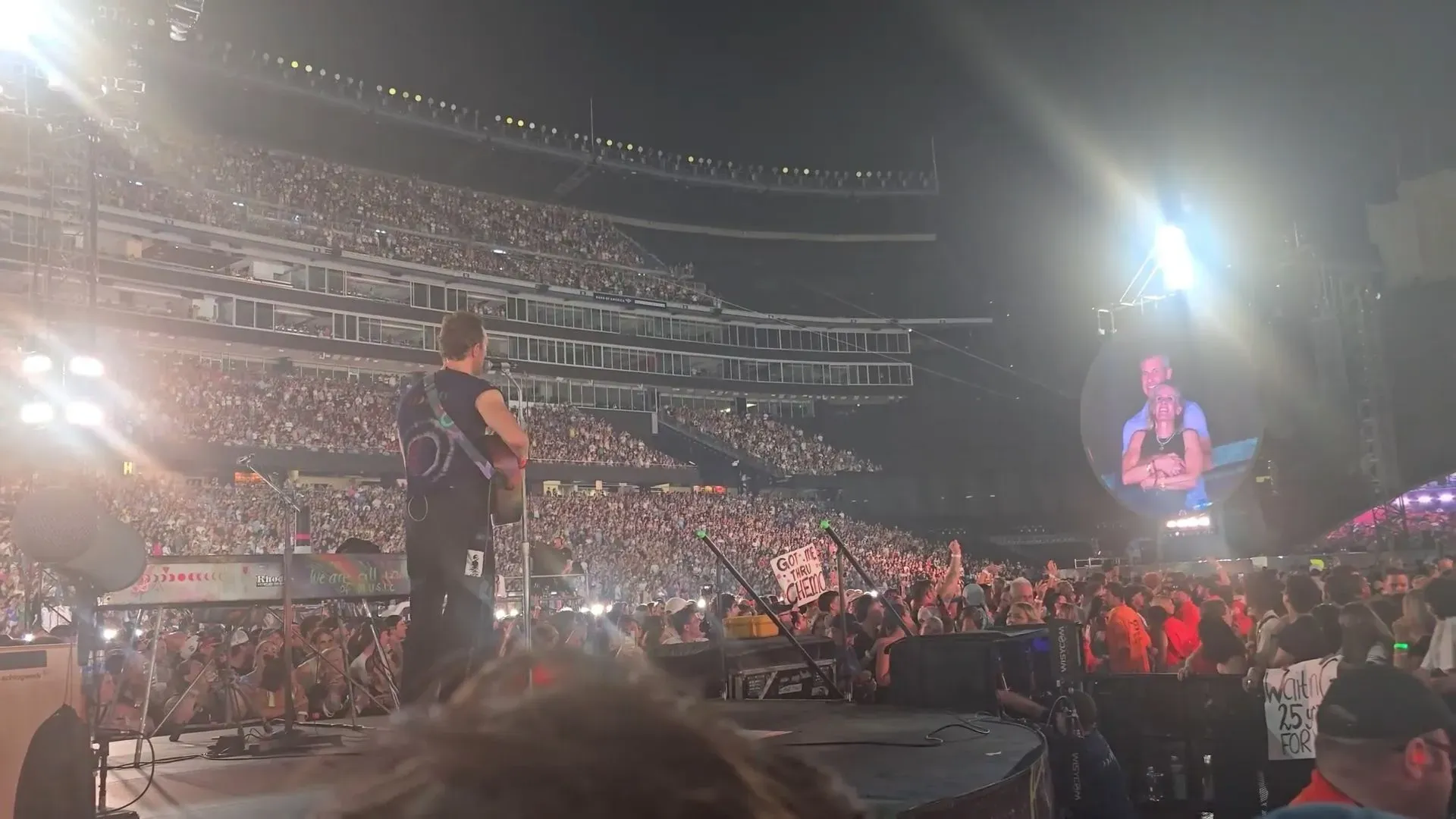 جمهور كبير يستمتع بحفل فرقة كولدبلاي في ملعب جيليت، مع ظهور كريستين كابوت وآندي بايرون على الشاشة الكبيرة.