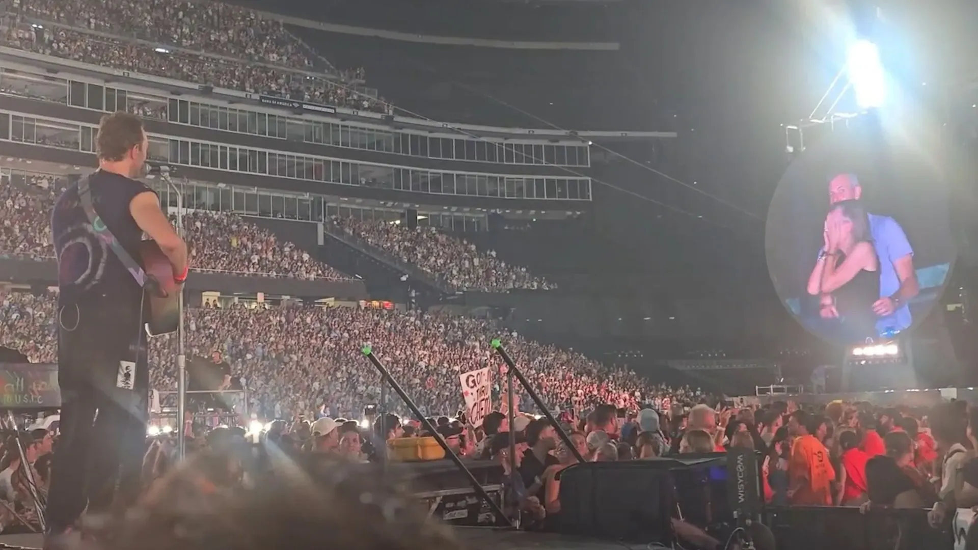 أندي بايرون وكريستين كابوت يحتضنان على شاشة عملاقة خلال حفل كولدبلاي في ملعب جيليت، وسط حشد جماهيري كبير.