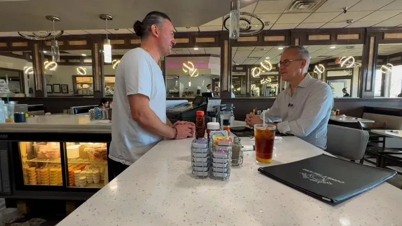 رجلان يتحدثان في مطعم، مع زجاجات صلصة وأكواب مشروبات على الطاولة، تعكس الأجواء الاقتصادية المتوترة في ميشيغان.