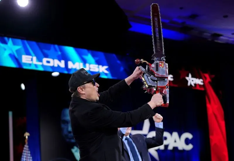إيلون ماسك يحمل منشارًا كهربائيًا في مؤتمر "CPAC"، مع خلفية تحمل اسمه، مما يعكس دعمه لترامب وعلاقته المتغيرة مع شركات التكنولوجيا.