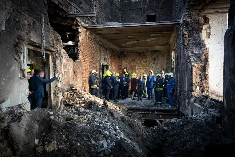 مجموعة من الأشخاص يرتدون خوذات واقية يتجمعون في موقع دمار، حيث تظهر الجدران المتضررة والأرض المليئة بالركام بعد الهجوم.