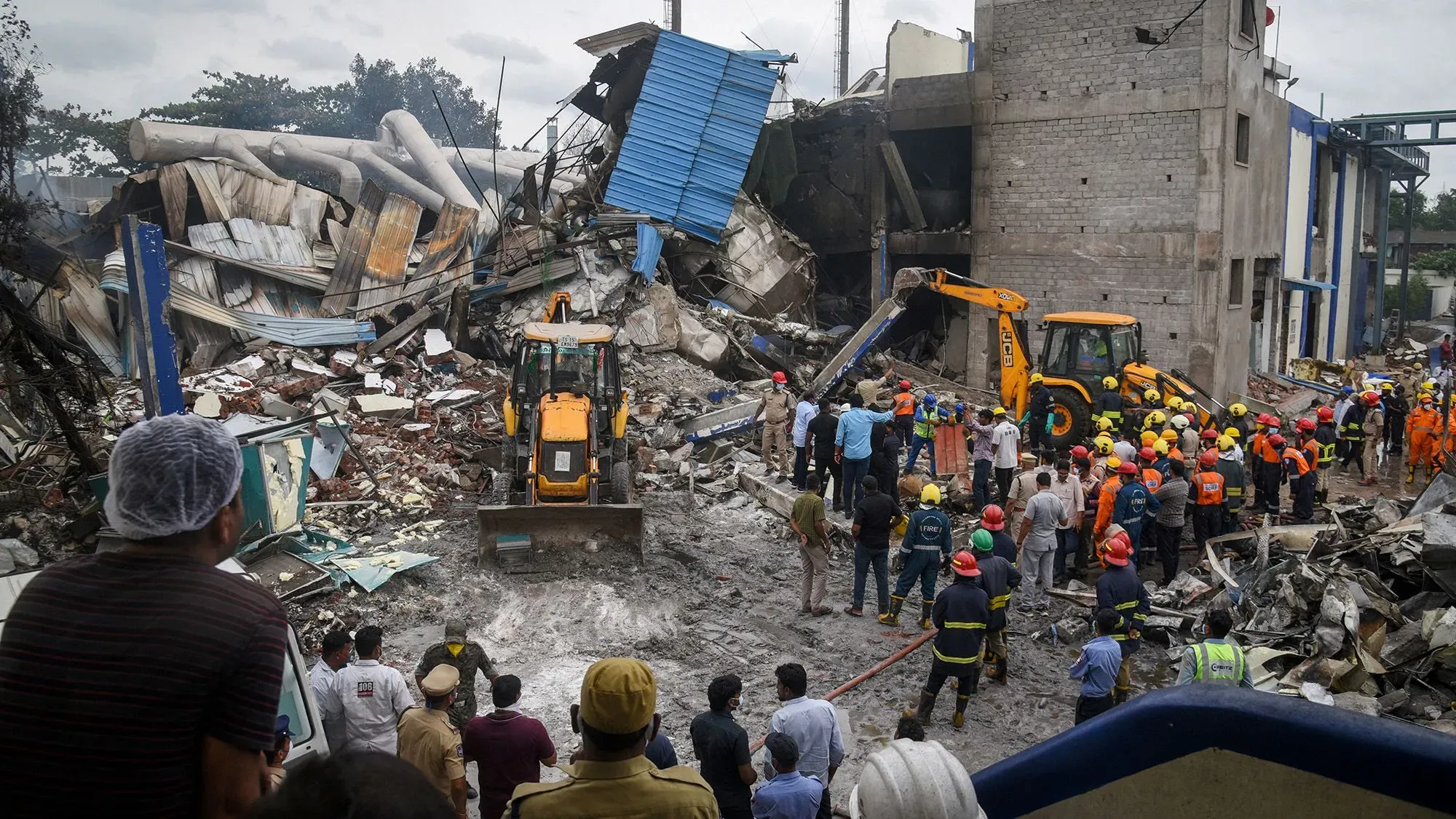 مشهد من موقع الانفجار في مصنع SIGC.NS في تيلانجانا، حيث يعمل رجال الإنقاذ وسط الحطام، مع وجود معدات ثقيلة وأشخاص يتجمعون لمراقبة جهود الإنقاذ.