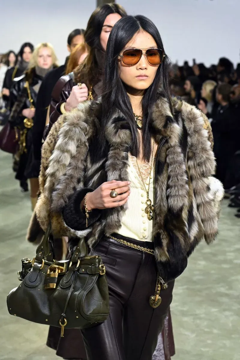 عارضة أزياء ترتدي معطفاً من الفرو مع نظارات شمسية، تحمل حقيبة يد، تسير على المدرج خلال عرض أزياء في أسبوع الموضة بباريس.