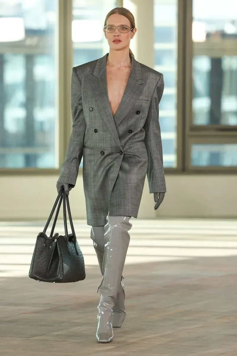 عارضة أزياء ترتدي سترة رمادية كبيرة مع بنطلون ضيق وحقيبة يد، تسير في عرض أزياء حديث بأجواء مكتبية، تعكس اتجاهات الموضة الجديدة.