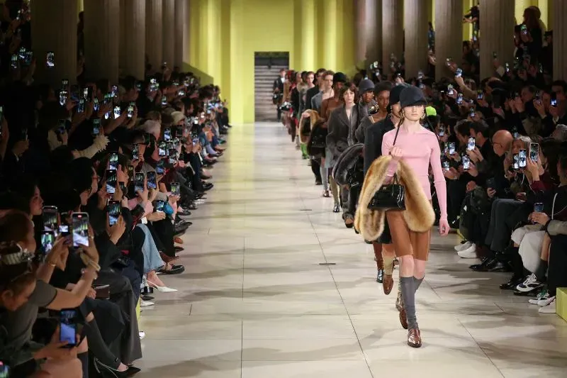 عرض أزياء في أسبوع الموضة بباريس، حيث تسير العارضات في ممر أمام جمهور يحمل هواتفهم، مع تصاميم متنوعة وأجواء حيوية.