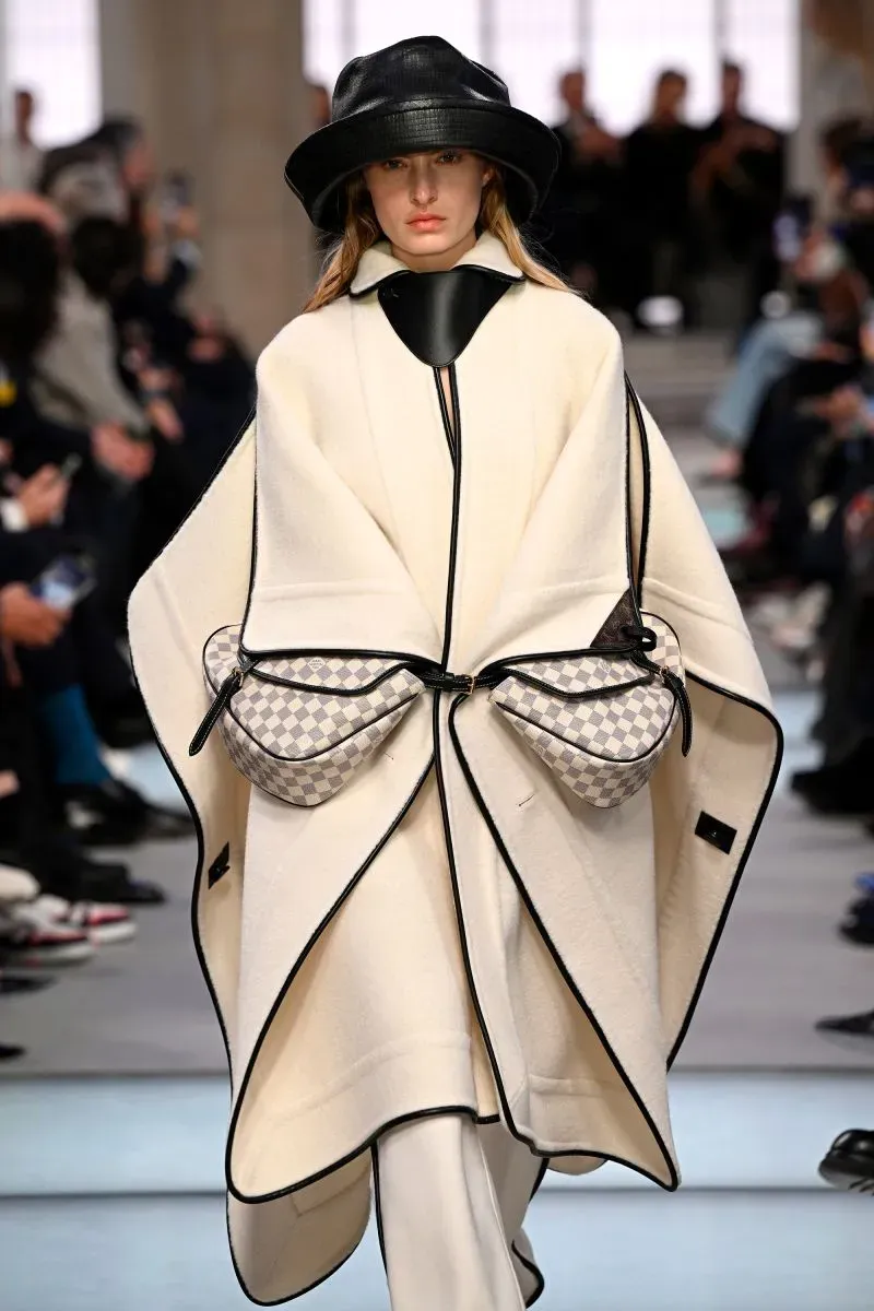 عارضة أزياء ترتدي عباءة بيضاء مع تفاصيل سوداء، تحمل حقيبتين مميزتين، خلال عرض أزياء في أسبوع الموضة بباريس.