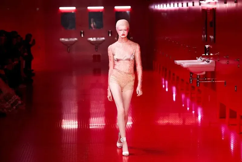 عارضة أزياء تسير في ممر أحمر مضاء، مرتدية ملابس داخلية من الدانتيل، مع خلفية تضم مغاسل وأضواء حمراء، تعكس أجواء عرض أزياء مبتكرة.