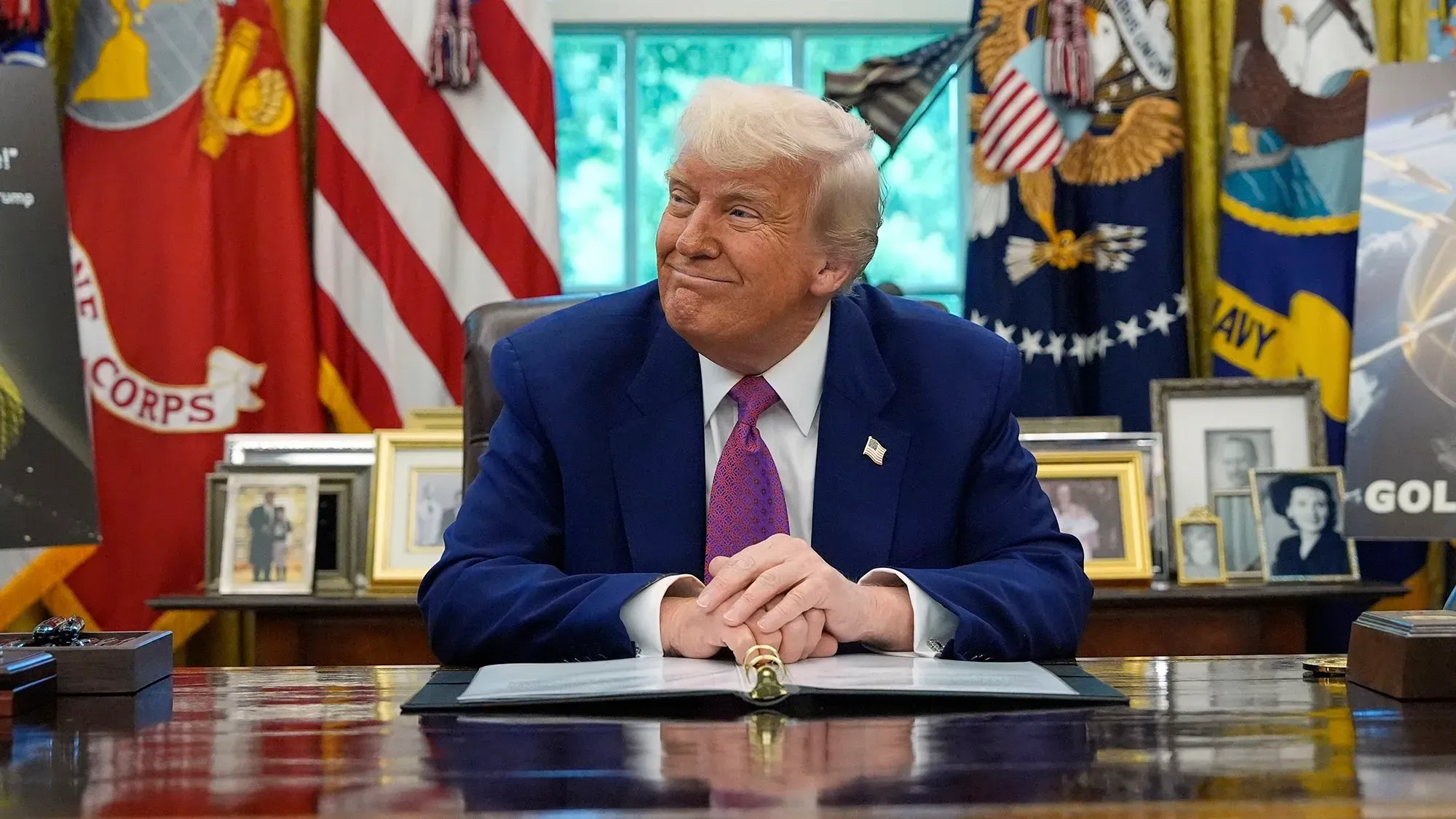 صورة لدونالد ترامب في المكتب البيضاوي، مبتسمًا أثناء توقيع مستندات، مع العلم الأمريكي والرموز الوطنية خلفه، تعكس أجواء حفل مزاد عملته المشفرة.