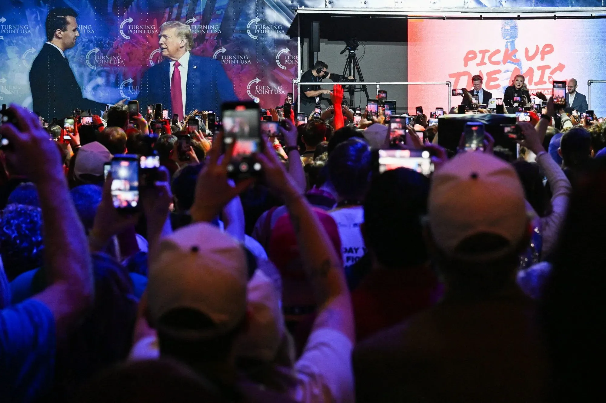 حشد كبير من الحضور في مهرجان أمريكا، يلتقطون الصور للرئيس السابق دونالد ترامب أثناء خطابه، مع لافتات Turning Point USA خلفهم.