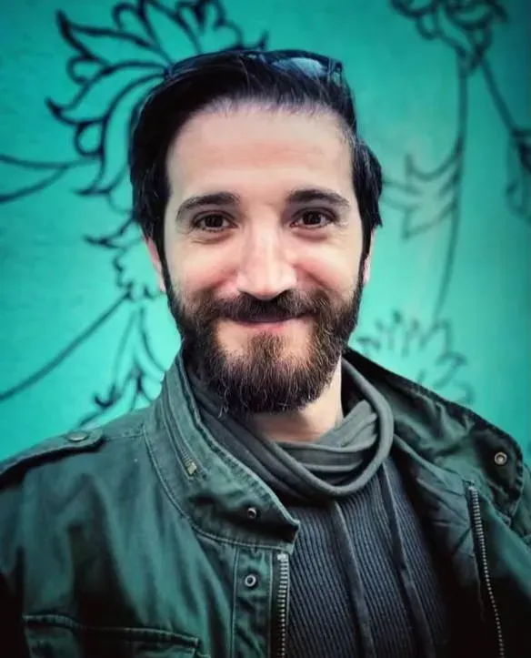 شخص مبتسم ذو لحية يرتدي سترة داكنة، يقف أمام جدار مزخرف بألوان زاهية، يعكس روح الحياة اليومية في طهران.