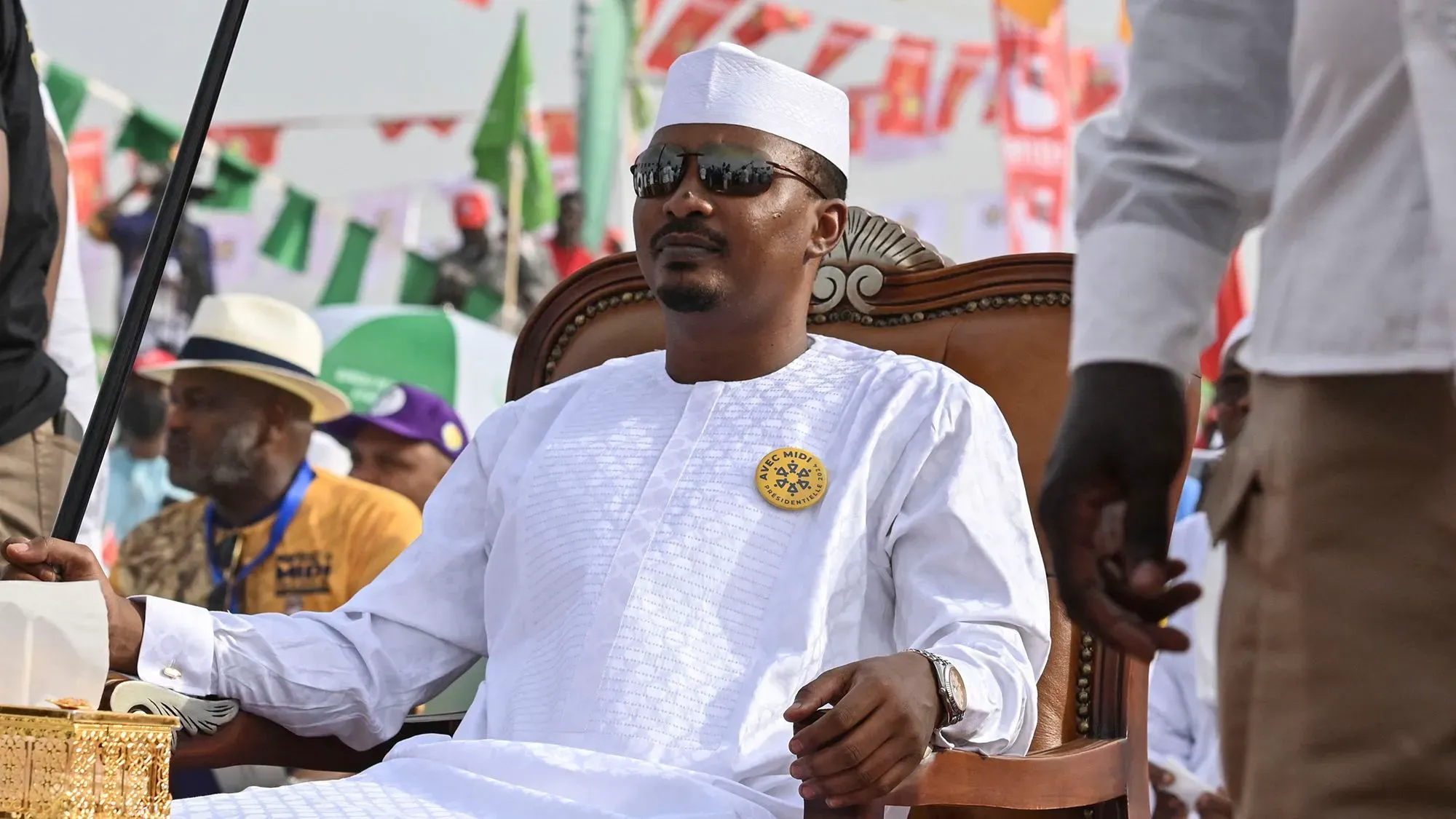 رجل يرتدي زيًا تقليديًا أبيض يجلس على كرسي مزخرف، محاط بجماهير في خلفية احتفالية. تعكس الصورة أجواء سياسية في تشاد.