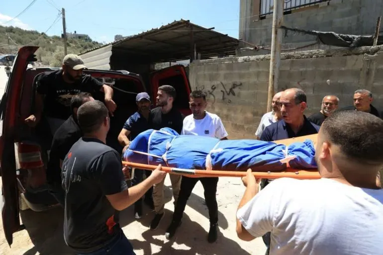 شبان يحملون جثمانًا مغطىً بقماش أزرق وسط مجموعة من الأشخاص في منطقة فلسطينية، في سياق تصاعد العنف ضد الفلسطينيين.