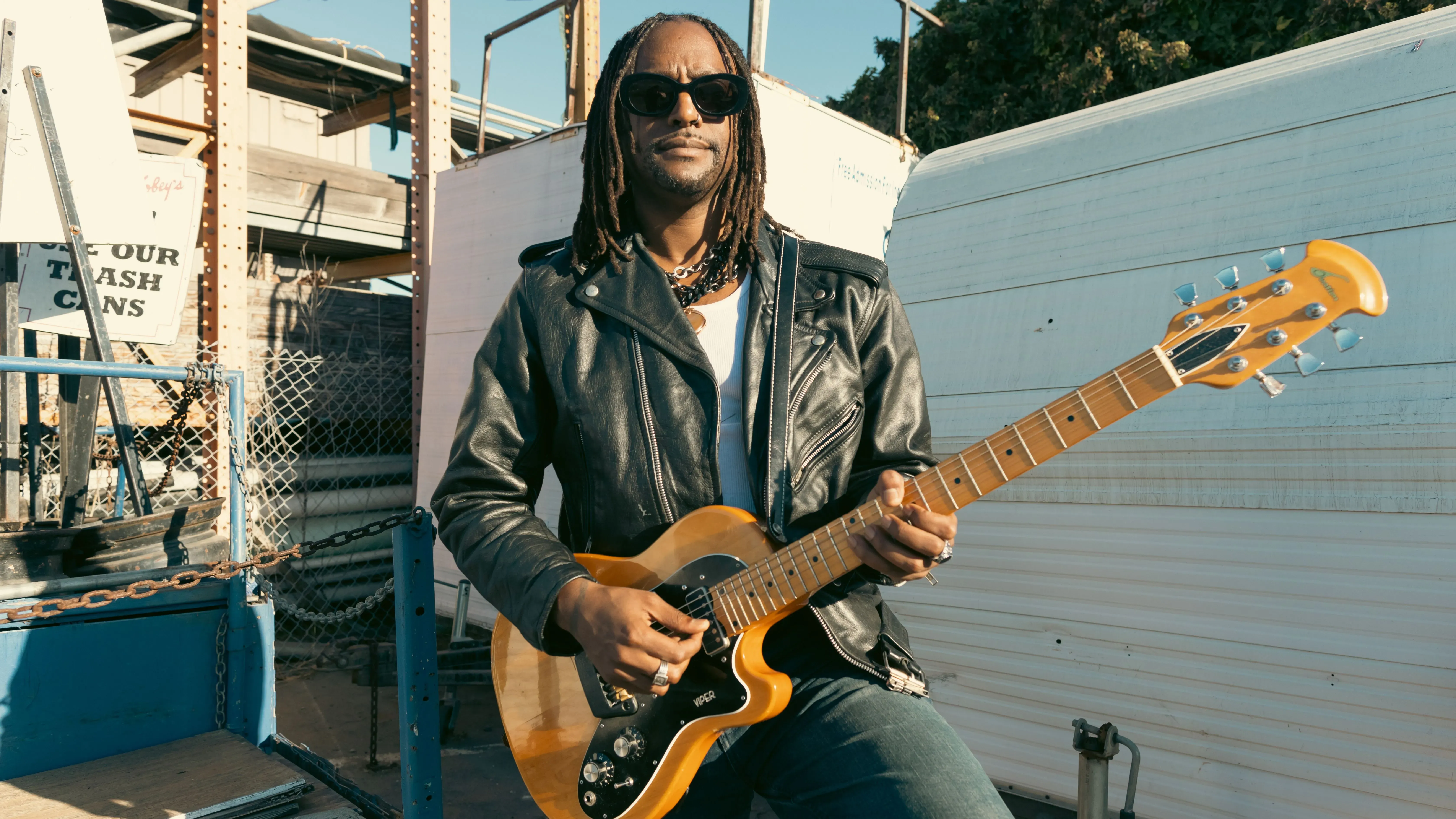 أوستن براون يعزف على الجيتار في موقع خارجي، مرتديًا سترة جلدية ونظارات شمسية، مع خلفية صناعية تعكس شغفه بالموسيقى.