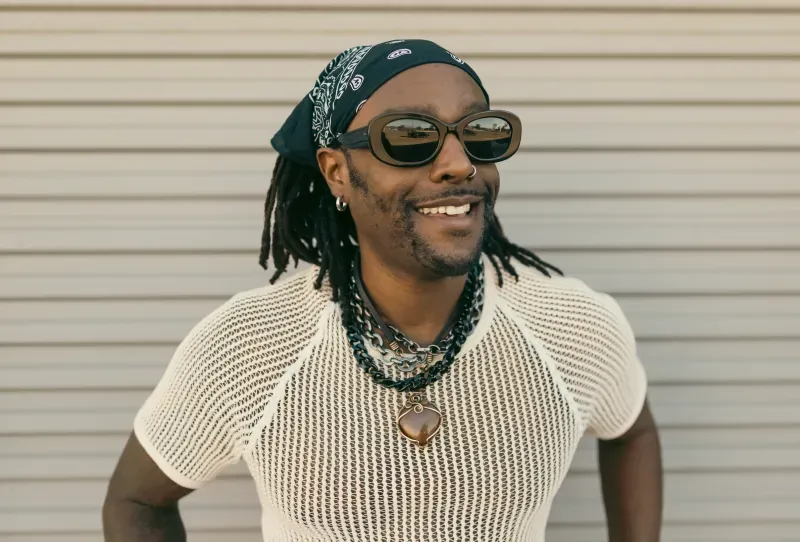 أوستن براون مبتسم ويرتدي نظارات شمسية وقميصًا شبكيًا، يقف أمام جدار رمادي، معبرًا عن شغفه بالموسيقى.