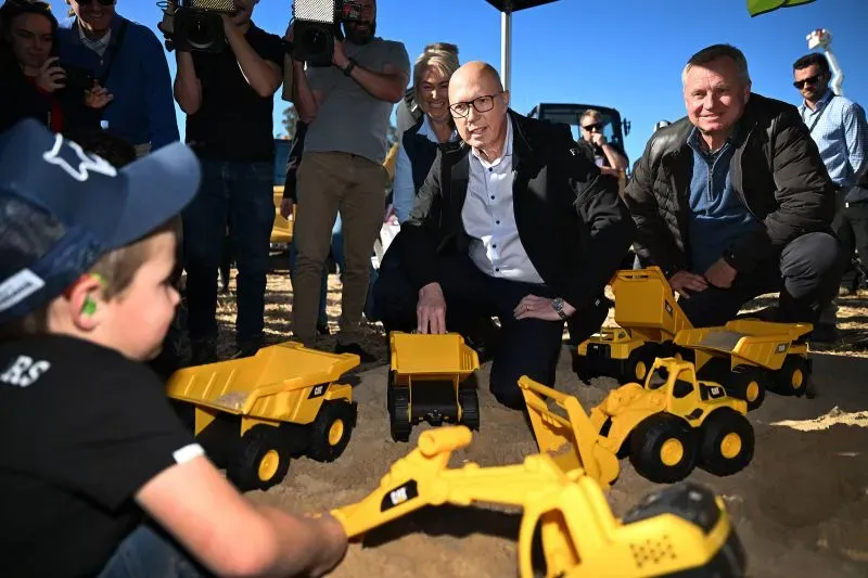 رجلان يجلسان بجانب طفل يلعب بمعدات بناء صغيرة على الرمال، مع وجود كاميرات وصحفيين في الخلفية، في سياق حملة انتخابية أسترالية.