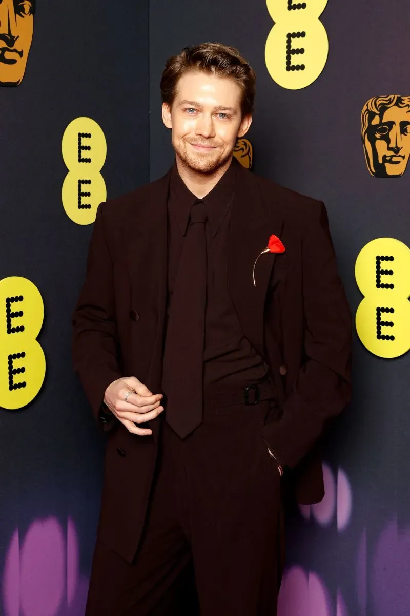 ممثل يرتدي بدلة سوداء مع وردة حمراء في حفل جوائز BAFTA، خلفية تحمل شعارات الحدث، يعكس أناقة الحفل.