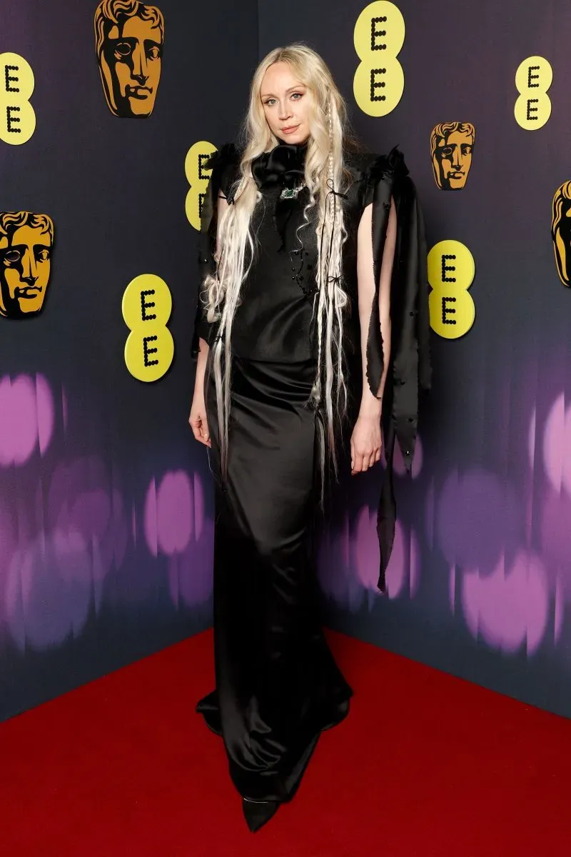 امرأة ترتدي فستاناً أسود أنيقاً مع تفاصيل مميزة، تقف على السجادة الحمراء لحفل جوائز BAFTA في لندن، مع خلفية تحمل شعارات الجائزة.