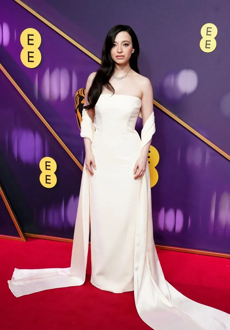ممثلة ترتدي فستاناً أبيض أنيقاً مع تفاصيل من الساتان، تقف على السجادة الحمراء لحفل جوائز BAFTA، مع خلفية ملونة.