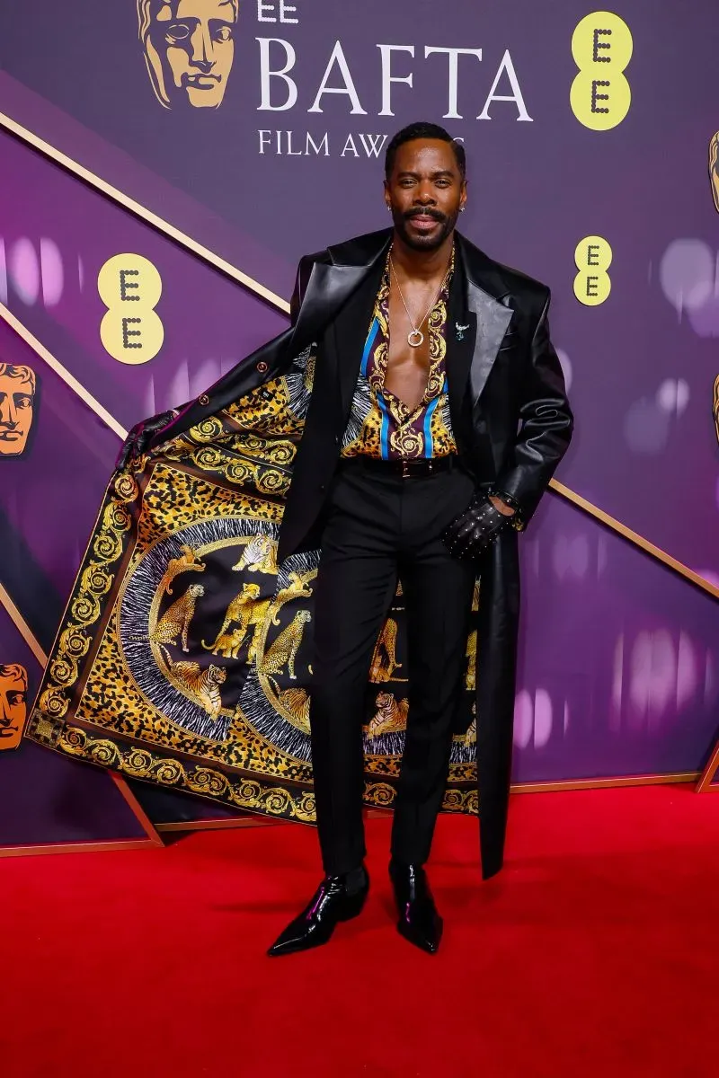 مظهر مميز لممثل يرتدي بدلة أنيقة مع قميص مزخرف، بينما يفتح معطفه في حفل جوائز BAFTA، مع خلفية ملونة تعكس أجواء الحدث.