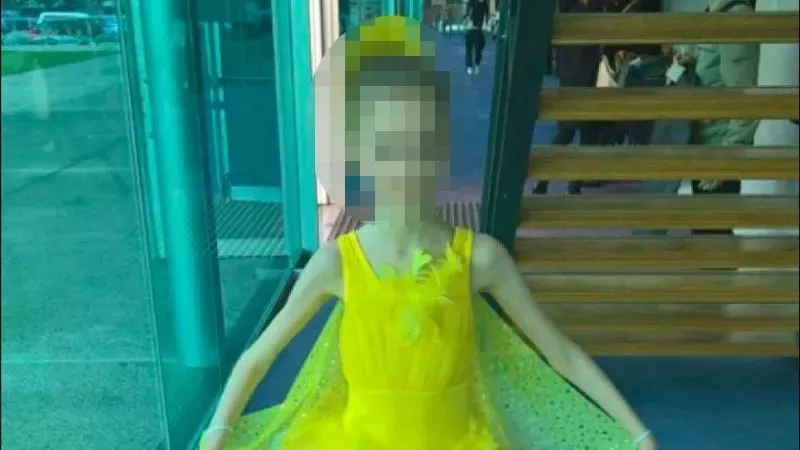 فتاة ترتدي فستاناً أصفر زاهياً، تظهر في وضعية رقص، تعكس براءة الطفولة، وسط خلفية معمارية حديثة.