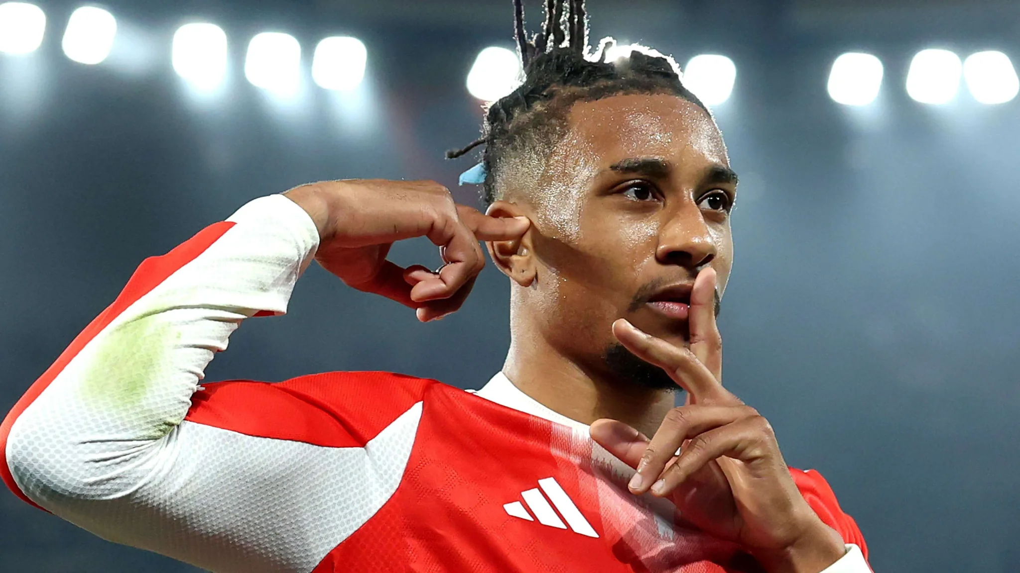 لاعب كرة قدم يحتفل بهدفه بإشارة الصمت، مرتديًا قميصًا أحمر وأبيض تحت أضواء ملعب مضيئة، في سياق سباق الكرة الذهبية 2026.