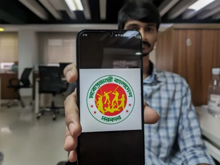 شخص يحمل هاتفًا ذكيًا يظهر شعار حكومة بنغلاديش، مع تعديل نص الشعار ليصبح "حكومة جمهورية بنغلاديش الغوغائية"، في سياق الاحتجاجات السياسية.