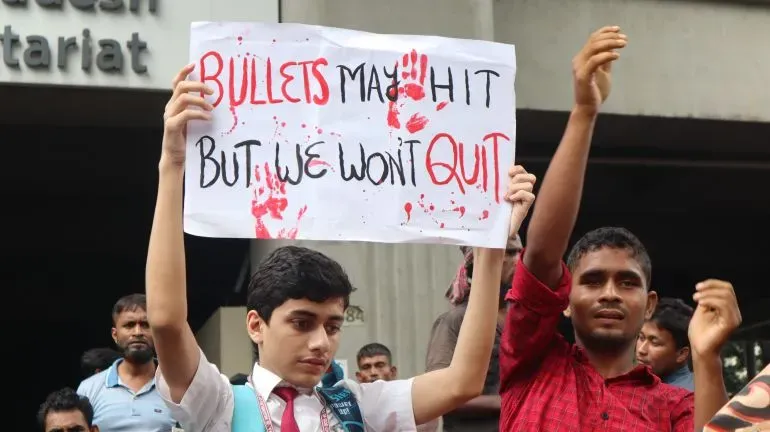 شاب يحمل لافتة مكتوب عليها "قد تصيبنا الرصاصات لكننا لن نستسلم" مع آثار دماء، وسط حشد من المتظاهرين في بنغلاديش.