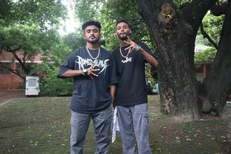 شخصان يرتديان ملابس عصرية، يقفان معًا في حديقة، بينما تظهر في الخلفية أشجار خضراء، مما يعكس ثقافة الشباب البنغلاديشي.