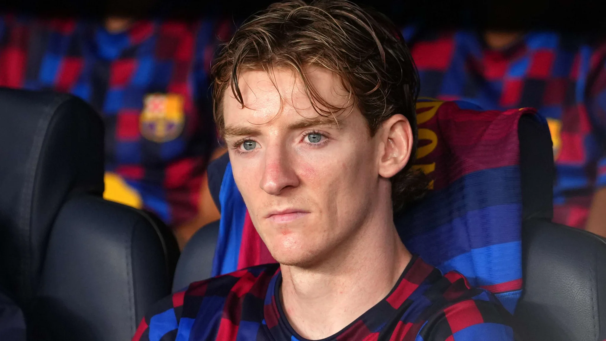 أنطوني جوردون لاعب جناح برشلونة مرتدٍ زي الفريق الرسمي، يظهر بتركيز وجدية على مقاعد البدلاء في ملعب كامب نو.