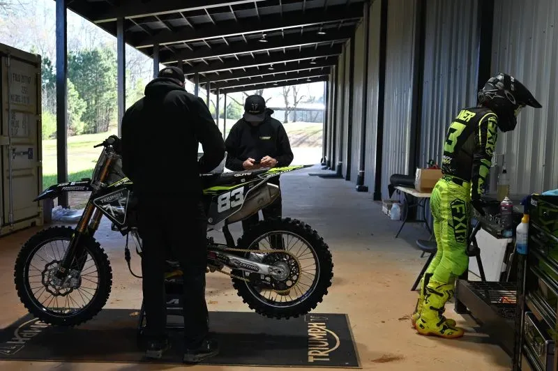 متسابقون في ورشة صيانة دراجات نارية، حيث يتم تجهيز الدراجة للسباق، مع التركيز على تفاصيل الأداء والتقنية العالية في رياضة السوبر كروس.