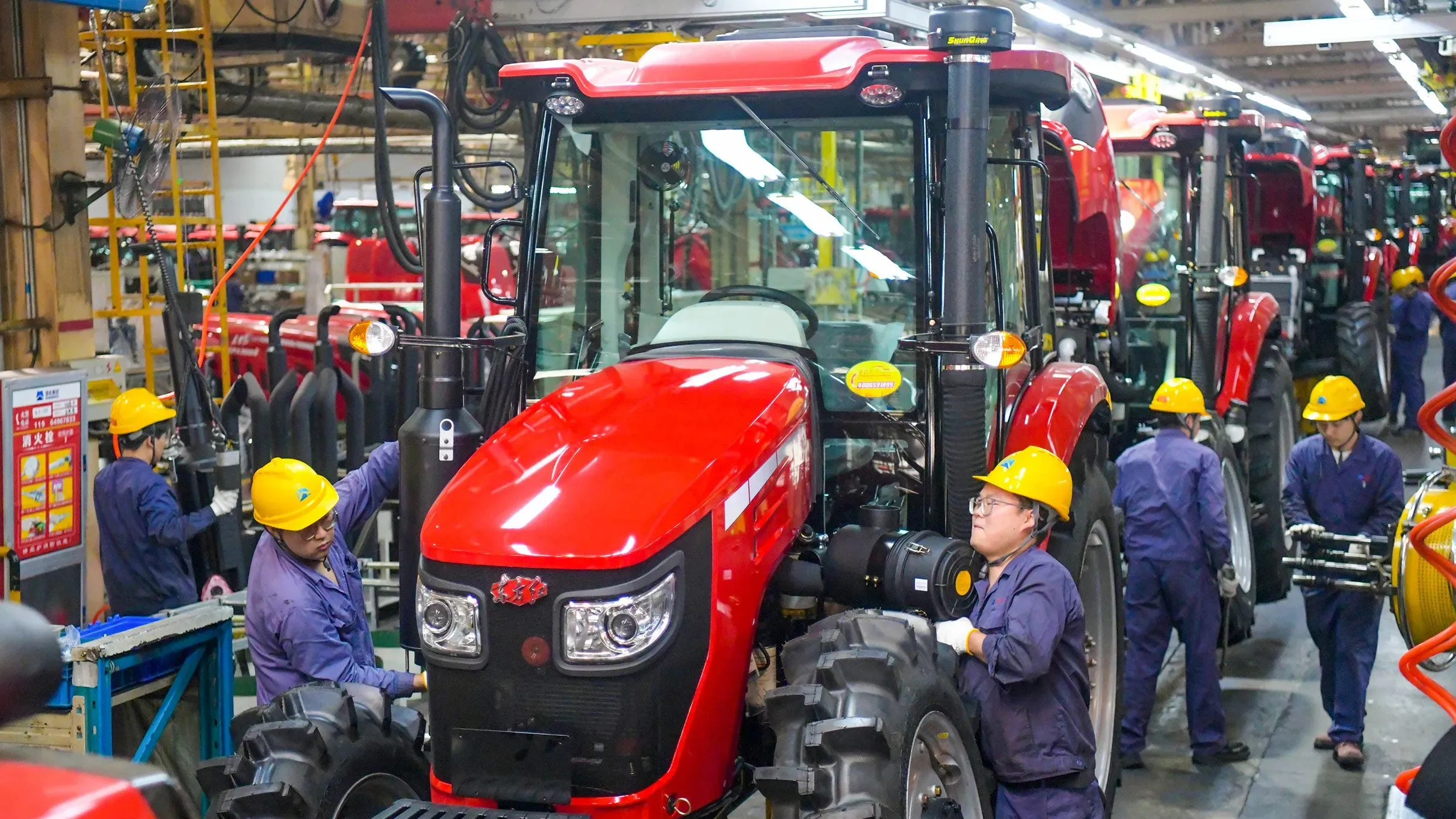 عمال يرتدون خوذات صفراء يعملون على تجميع جرارات حمراء في مصنع بالصين، في سياق التوترات التجارية بين الولايات المتحدة والصين.