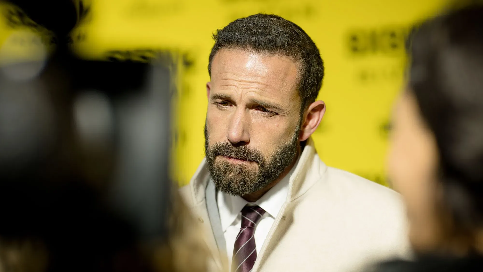 بين أفليك يظهر في حدث مع خلفية صفراء، يعكس تعابير وجهه الجادة. يتناول مواضيع حياته الشخصية والمهنية في مقابلة مع مجلة GQ.