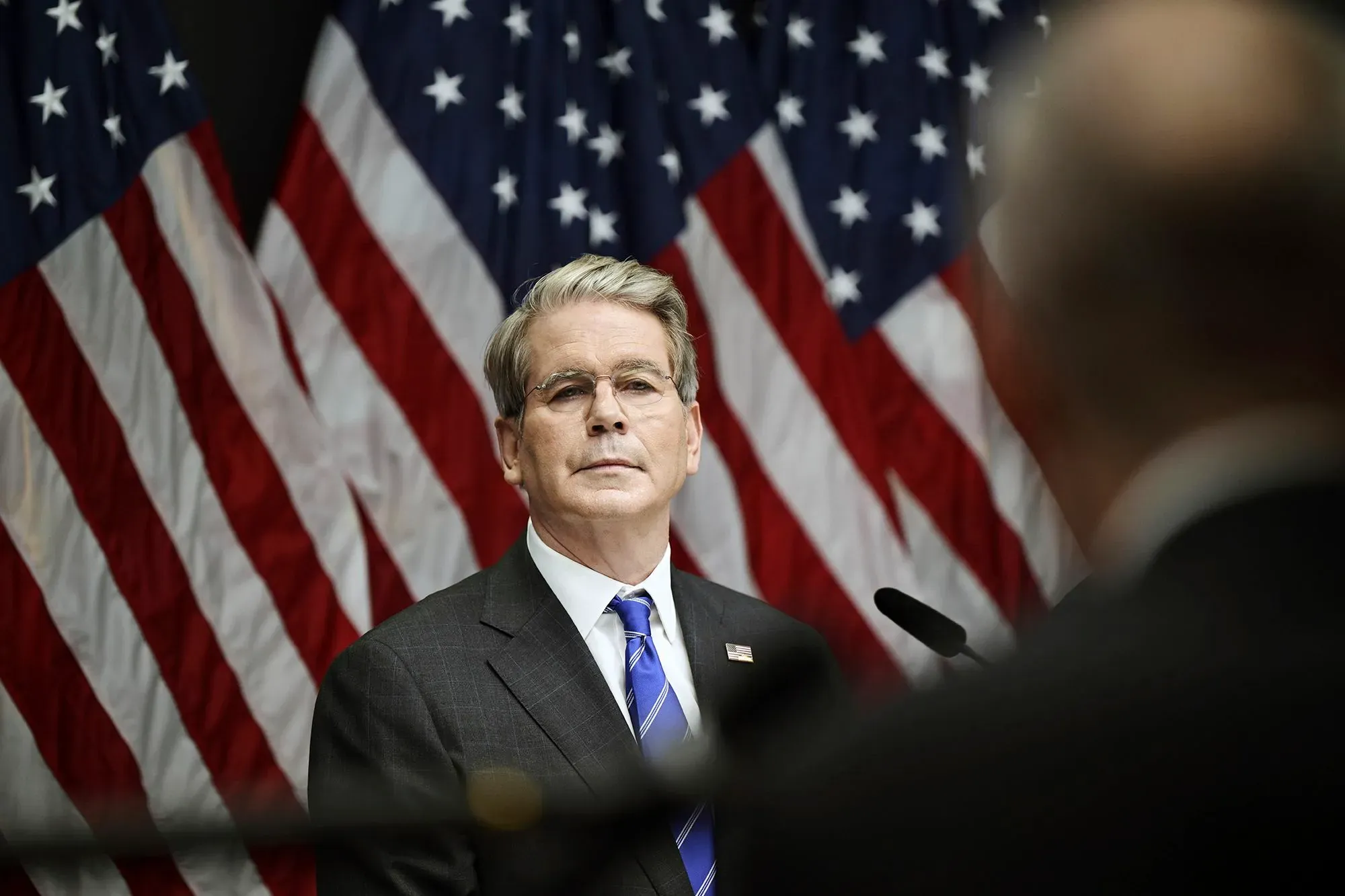 وزير الخزانة سكوت بيسنت يتحدث خلف ميكروفون، مع أعلام أمريكية خلفه، في سياق مناقشة تنظيم البنوك.