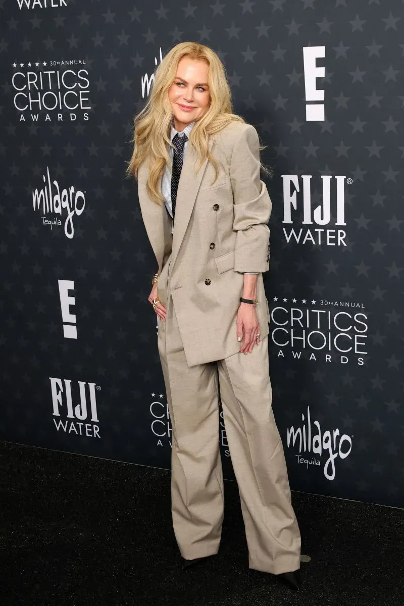 نيكول كيدمان ترتدي بدلة أنيقة بلون البيج في حفل توزيع جوائز اختيار النقاد، مع خلفية تحمل شعارات الحفل والمشروبات.