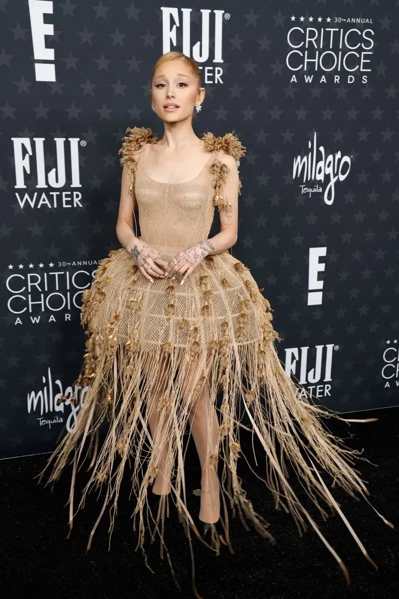 تظهر امرأة ترتدي فستانًا مبتكرًا مزينًا بخيوط ذهبية، في حفل توزيع جوائز اختيار النقاد، مع خلفية تحمل شعارات الحدث.