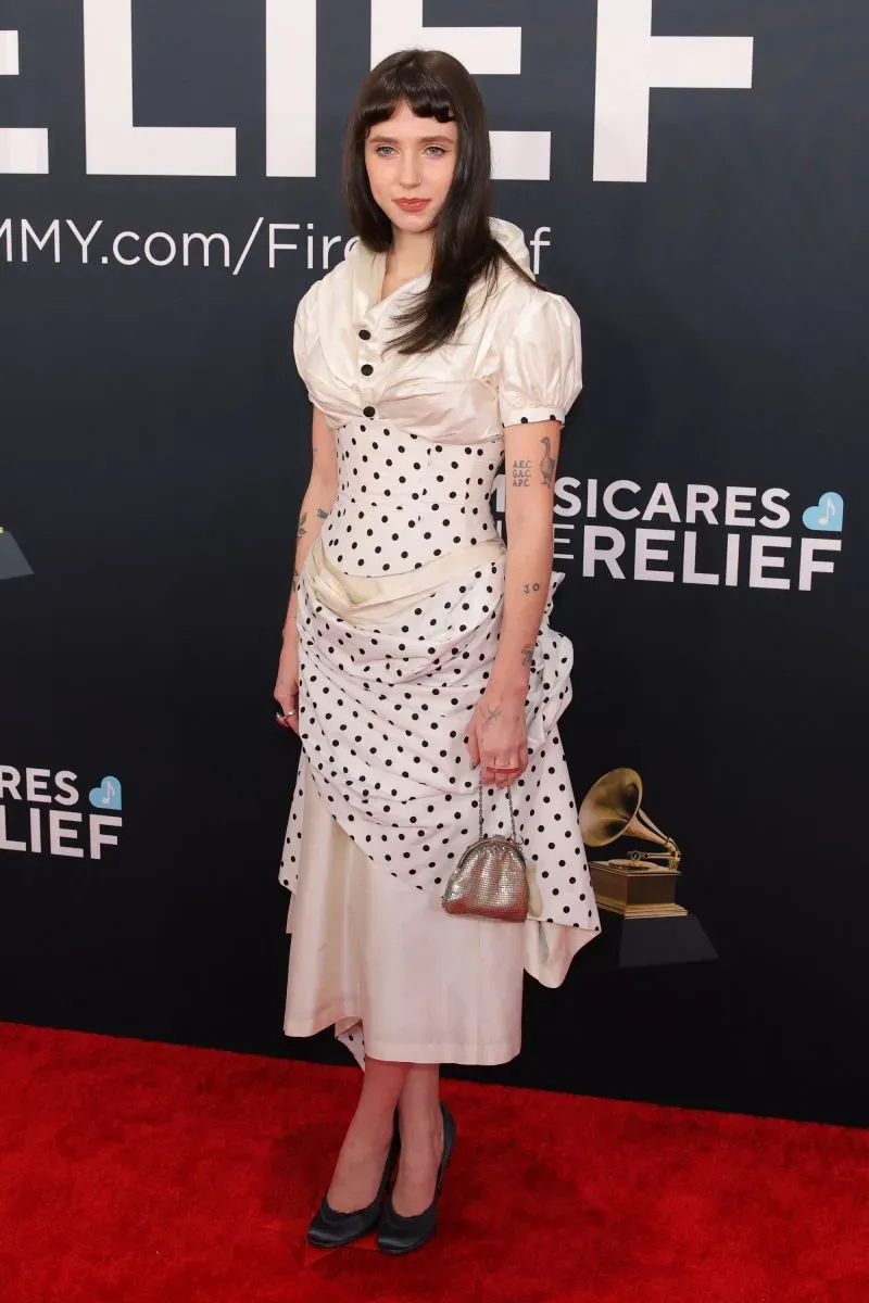 شخصية ترتدي فستانًا أبيض مزين بنقاط سوداء، تحمل حقيبة صغيرة، في حفل توزيع جوائز غرامي على السجادة الحمراء.