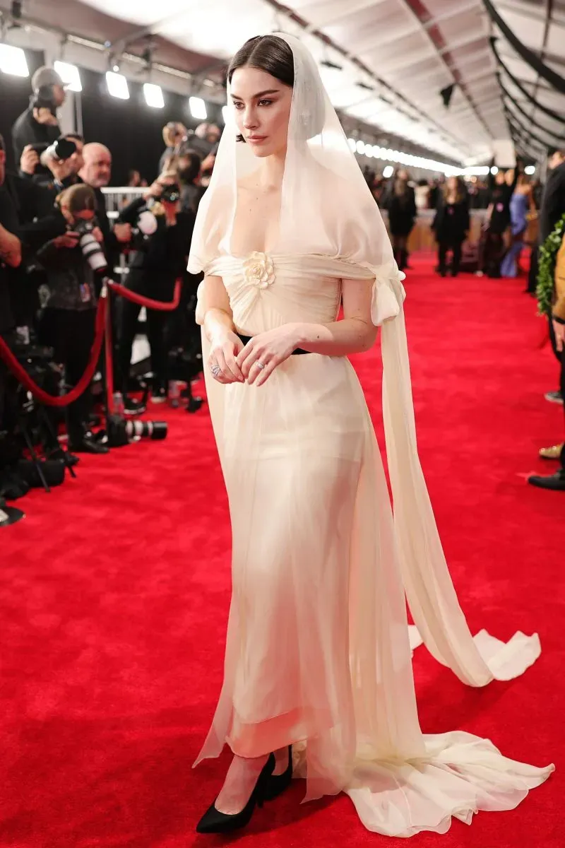 مشاركة فنانة ترتدي فستانًا أبيض شفافًا مع غطاء رأس، تسير على السجادة الحمراء في حفل توزيع جوائز غرامي، محاطة بكاميرات المصورين.