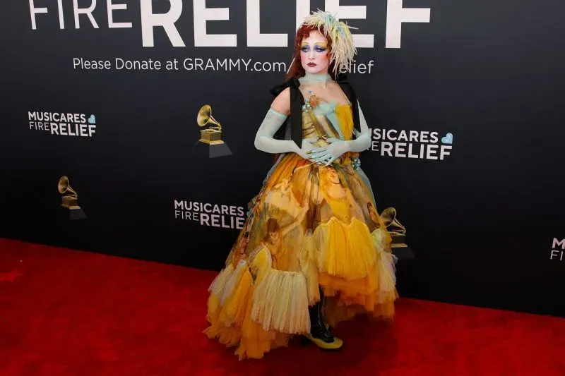 شخص يرتدي فستانًا ملونًا ومزخرفًا، يقف على السجادة الحمراء خلال حفل توزيع جوائز غرامي، مع خلفية تشير إلى دعم جهود الإغاثة من حرائق الغابات.