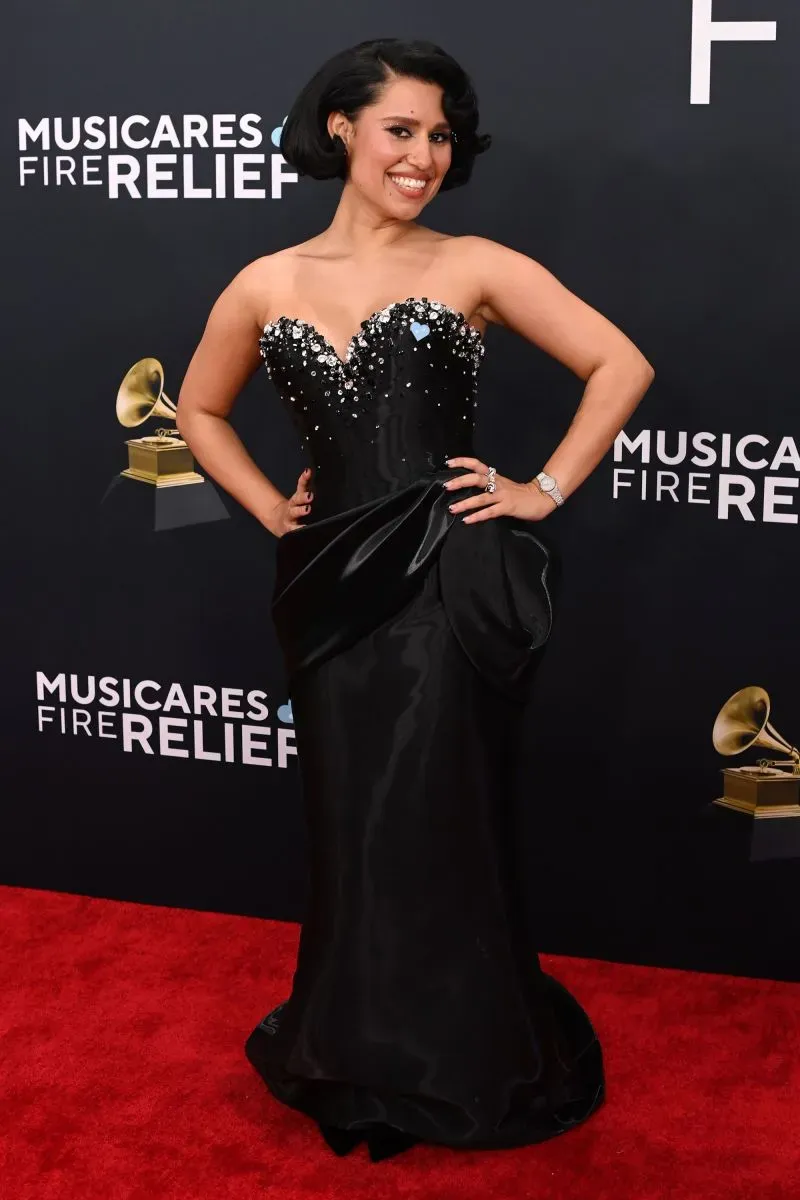 امرأة ترتدي فستانًا أسود مزينًا بالخرز اللامع، تبتسم على السجادة الحمراء لحفل توزيع جوائز غرامي، مع شعار "موسيكيرز للإغاثة من الحرائق" في الخلفية.